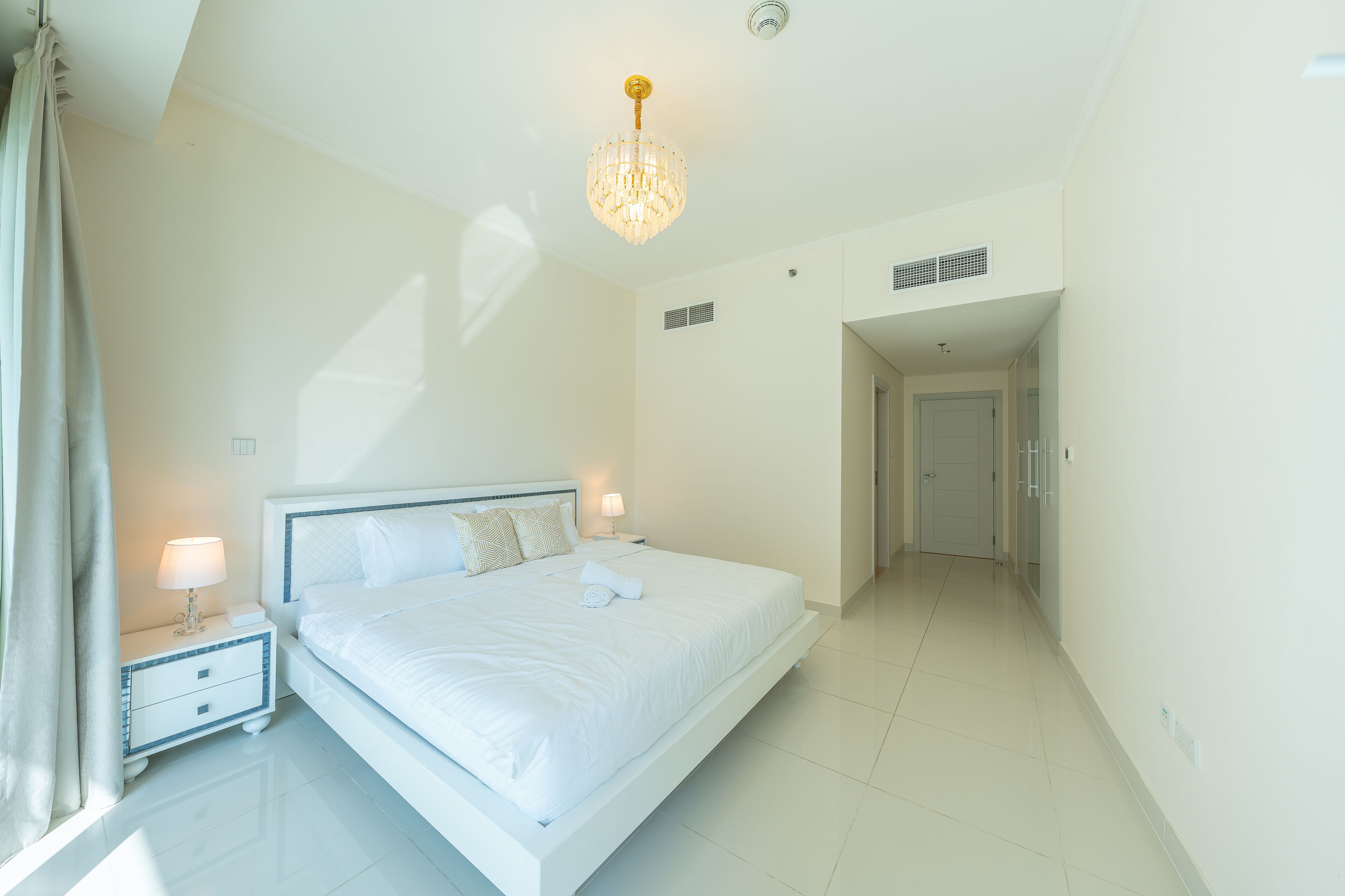 A modern hotel room with a large bed, bedside tables, a chandelier, and a view of the hallway.