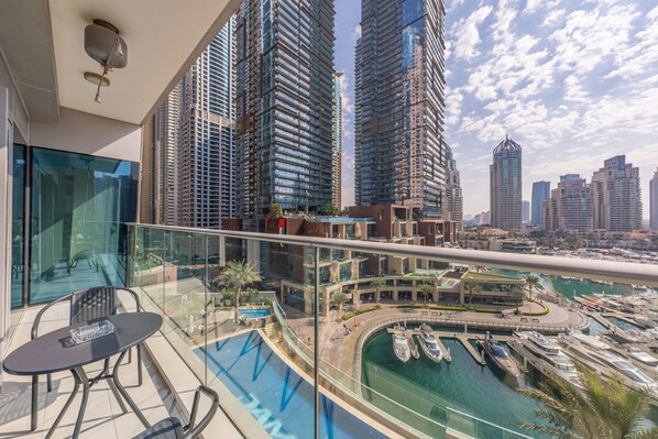 A balcony with a glass railing overlooking a marina with boats, a pool, and tall buildings in the background.