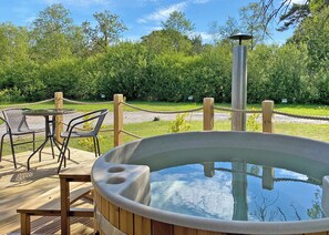 Un jacuzzi en una terraza de madera con sillas y una barandilla de cuerda, rodeado de follaje.