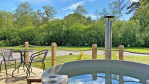 A hot tub on a wooden deck with chairs and a rope barrier, surrounded by greenery. - Pet Friendly Rustic Pod (Hale)