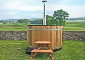 Una sauna de madera con chimenea, un banco y una mesa, ubicada en un entorno exterior con césped y vistas a un paisaje montañoso.
