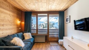 A cozy room with a wooden ceiling, a dark blue sofa with fluffy pillows, a white dresser, and a large window offering a view of snow-capped mountains. - Le Curling A (Val Claret) by Interhome (Tignes)