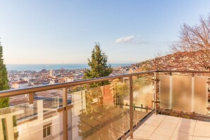 Un balcón con barandilla de vidrio, vista panorámica de una ciudad costera y cielo despejado.