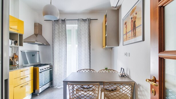 Een kleine keuken met een fornuis, gele kastjes en een eethoek met een tafel en stoelen.