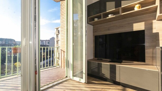 Un balcón con puerta corredera de vidrio, un televisor de pantalla plana montado en un mueble de madera y vista panorámica de la ciudad.