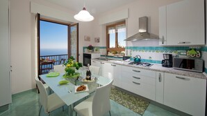Eine moderne Küche mit weißen Schränken, einem Esstisch mit Stühlen, Blick auf das Meer durch eine offene Tür und ein Balkon mit Tisch und Stühlen.