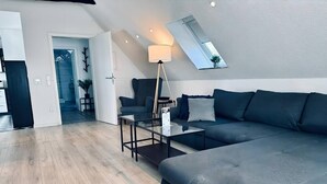 Un salon moderne avec un grand canapé gris, une table basse en verre et un lampadaire. Une lucarne se trouve au-dessus du canapé, offrant une vue sur l’extérieur.