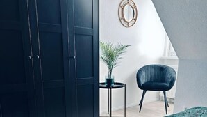 Une chambre avec une grande armoire, une petite table ronde ornée d’une plante, un fauteuil sombre et une couvre-lit à motifs.