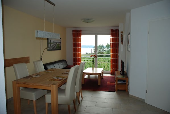 Ein Essbereich mit einem Holztisch, weißen Stühlen, einer Bank und Blick auf Grünflächen und ein Gewässer durch große Fenster.
