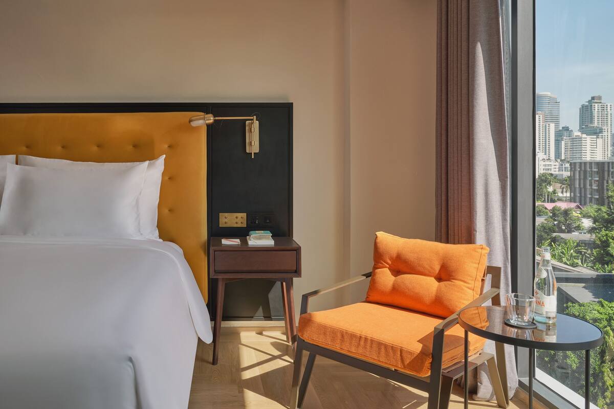 A hotel room with a bed, a nightstand, a chair, a table, and a city view through large windows.