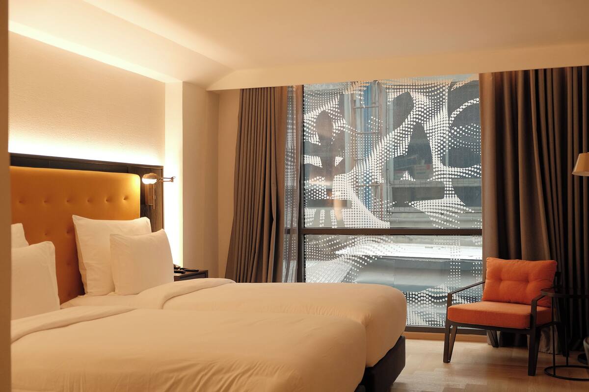 A hotel room with a large bed, a nightstand, a chair, and a window with a view of a cityscape.