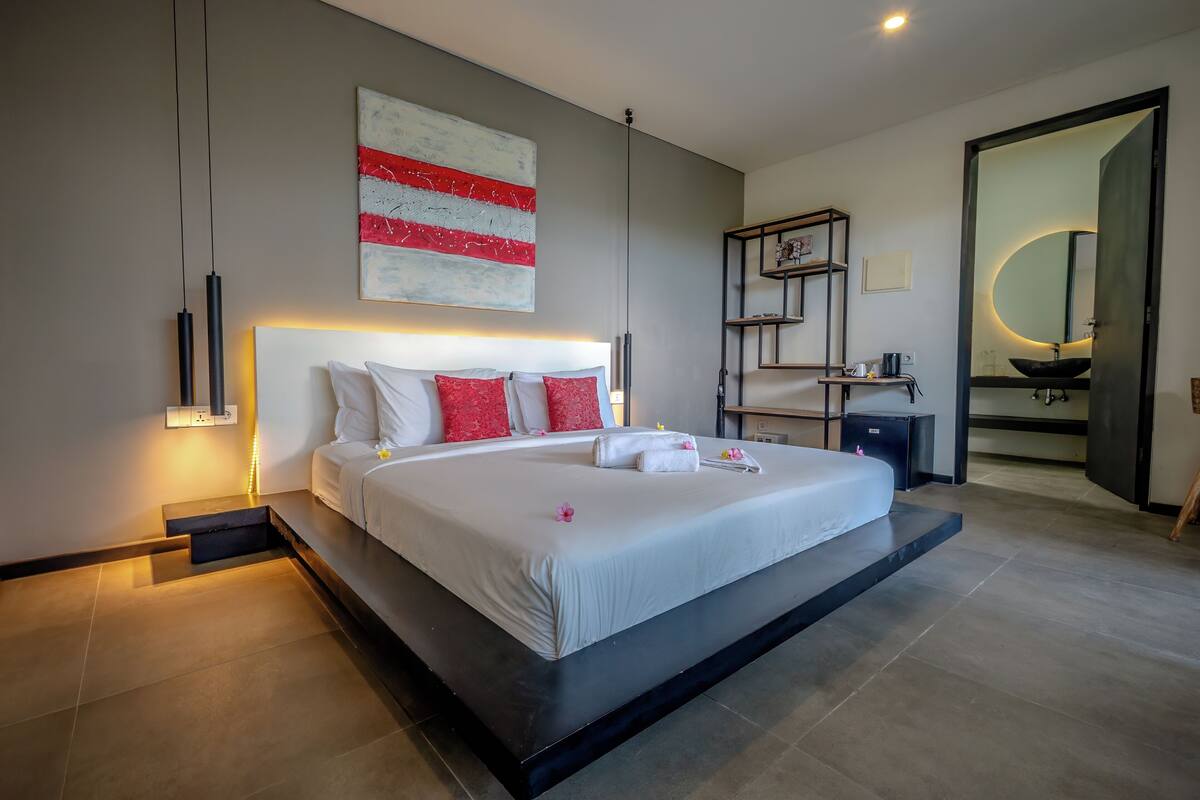 Una habitación de hotel moderna con una cama grande, mesitas de noche, una lámpara de noche, una obra de arte colgada en la pared, un estante empotrado y un baño con lavabo y espejo.