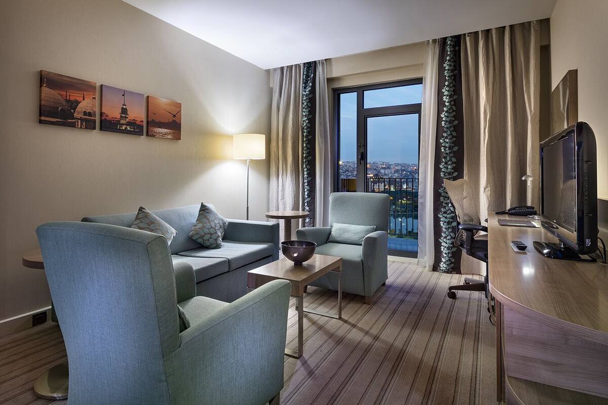 Une chambre d’hôtel équipée d’un canapé, de fauteuils, d’un bureau, d’une télévision et offrant une vue sur le paysage urbain.
