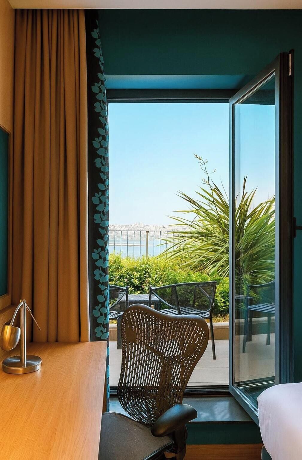 Ein Hotelzimmer mit einer großen Schiebetür, die auf einen Balkon mit Blick auf eine Stadtlandschaft und Grünflächen führt.