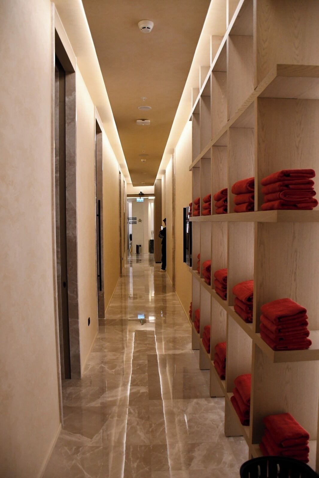 Un couloir d’hôtel avec un sol en marbre, des étagères en bois où sont pliées des serviettes rouges, et un design épuré et moderne.