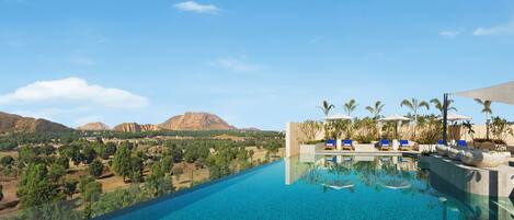 A pool with clear blue water, surrounded by lounge chairs and umbrellas, with a view of mountains and greenery in the background.