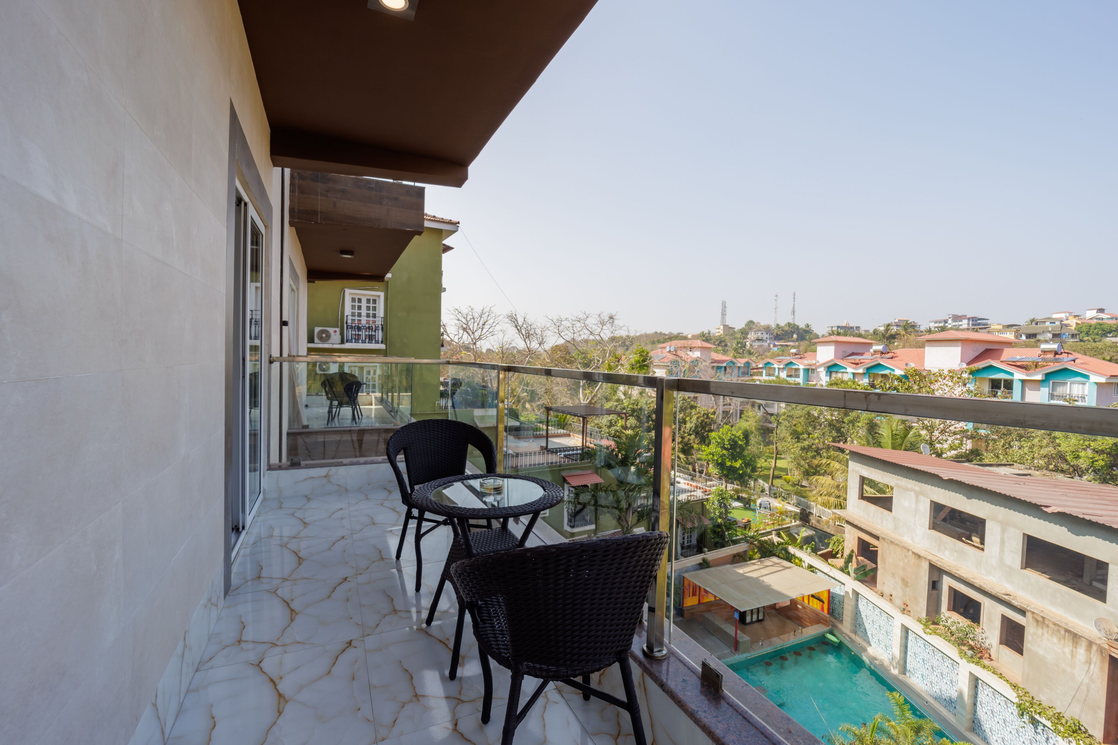 Un balcón con barandilla de vidrio, dos sillas de mimbre, una mesa redonda pequeña y vistas a una piscina y edificios al fondo.