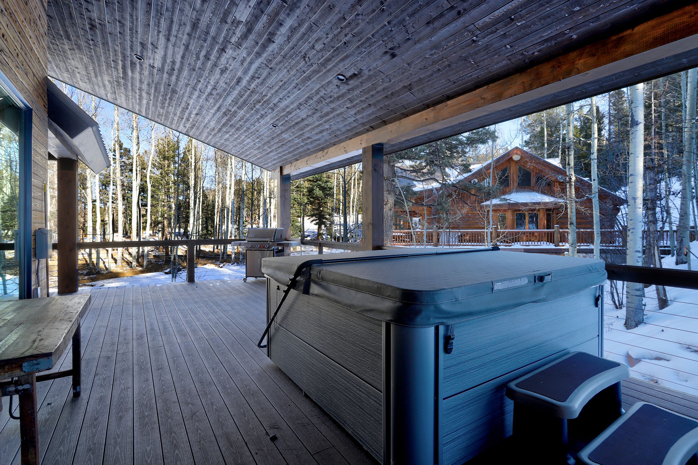 A wooden deck with a hot tub, a table, and a view of a cabin in the background.