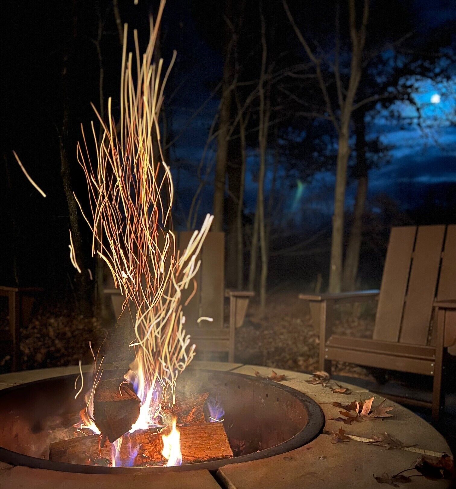 A fire pit with burning logs and sparks, surrounded by chairs and a night sky.