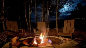 A circular fire pit with logs, surrounded by wooden chairs, set in a wooded area at night. - Moonlight View @ Spring Brook Resort (Wisconsin Dells)