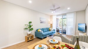 A modern living room with a blue sofa, a dining table set with plates and a bottle, and a TV mounted on the wall. - 5 Gretel Lodge, 1 Gretel Cl - family friendly unit close to the water (Nelson Bay)