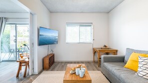 Un salon moderne avec un téléviseur à écran plat, un canapé gris, une table basse en bois sur laquelle sont disposées des pâtisseries, et une petite table d’appoint en bois.
