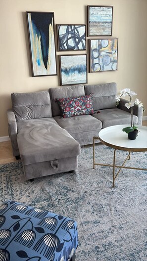 Un salon moderne avec un canapé d’angle gris, un pouf à motifs, une table basse ronde ornée d’une orchidée en pot et des œuvres d’art abstraites accrochées au mur.
