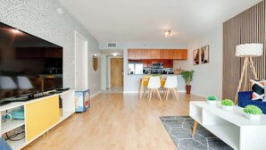 A modern living room with a flat-screen TV, wooden flooring, a dining area with chairs, a kitchen in the background, and a white console table with decorative plants. - Ocean&City Views Lux Miami Brickell Sleeps 4 KngBd (Miami)