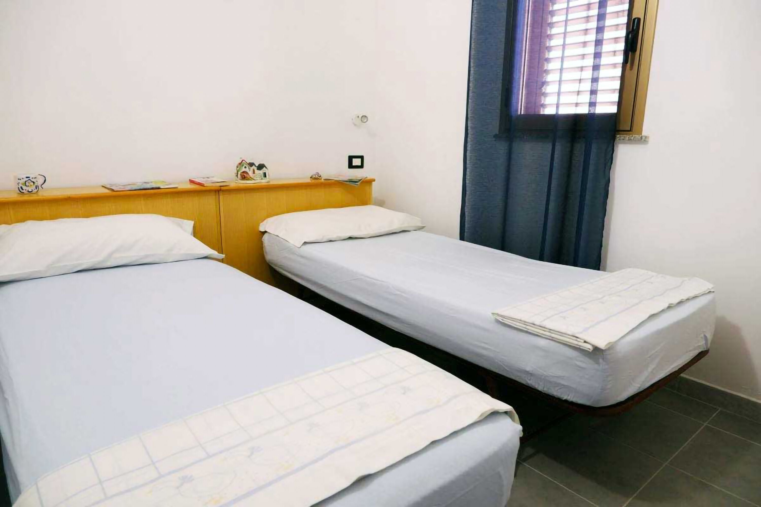 Een kamer met twee eenpersoonsbedden, een houten hoofdbord, een klein tafeltje met een klok en een raam met zonwering.