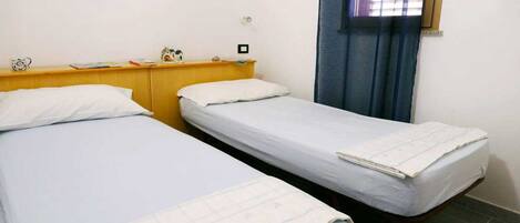 A room with two single beds, a wooden headboard, a small table with a clock, and a window with blinds.