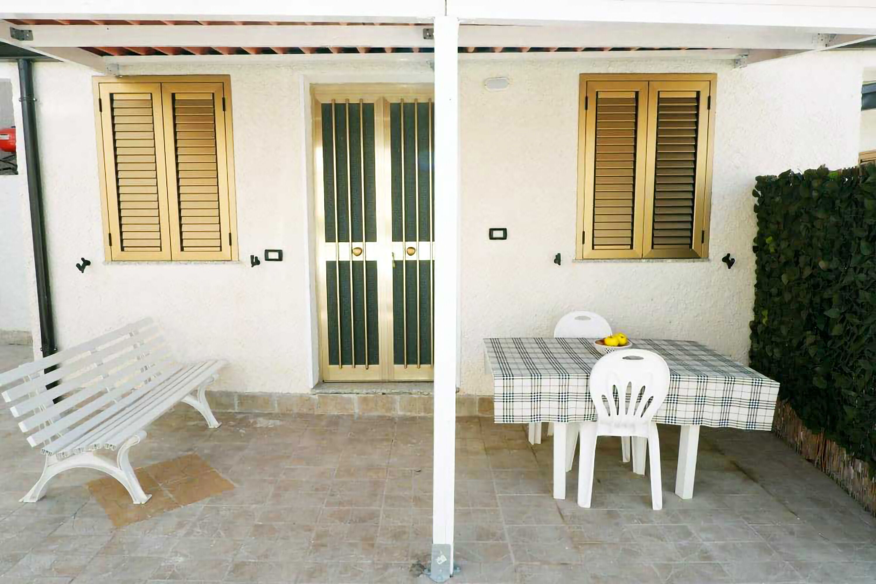 A patio area with a white bench, a table with a checkered tablecloth, and two windows with wooden shutters.