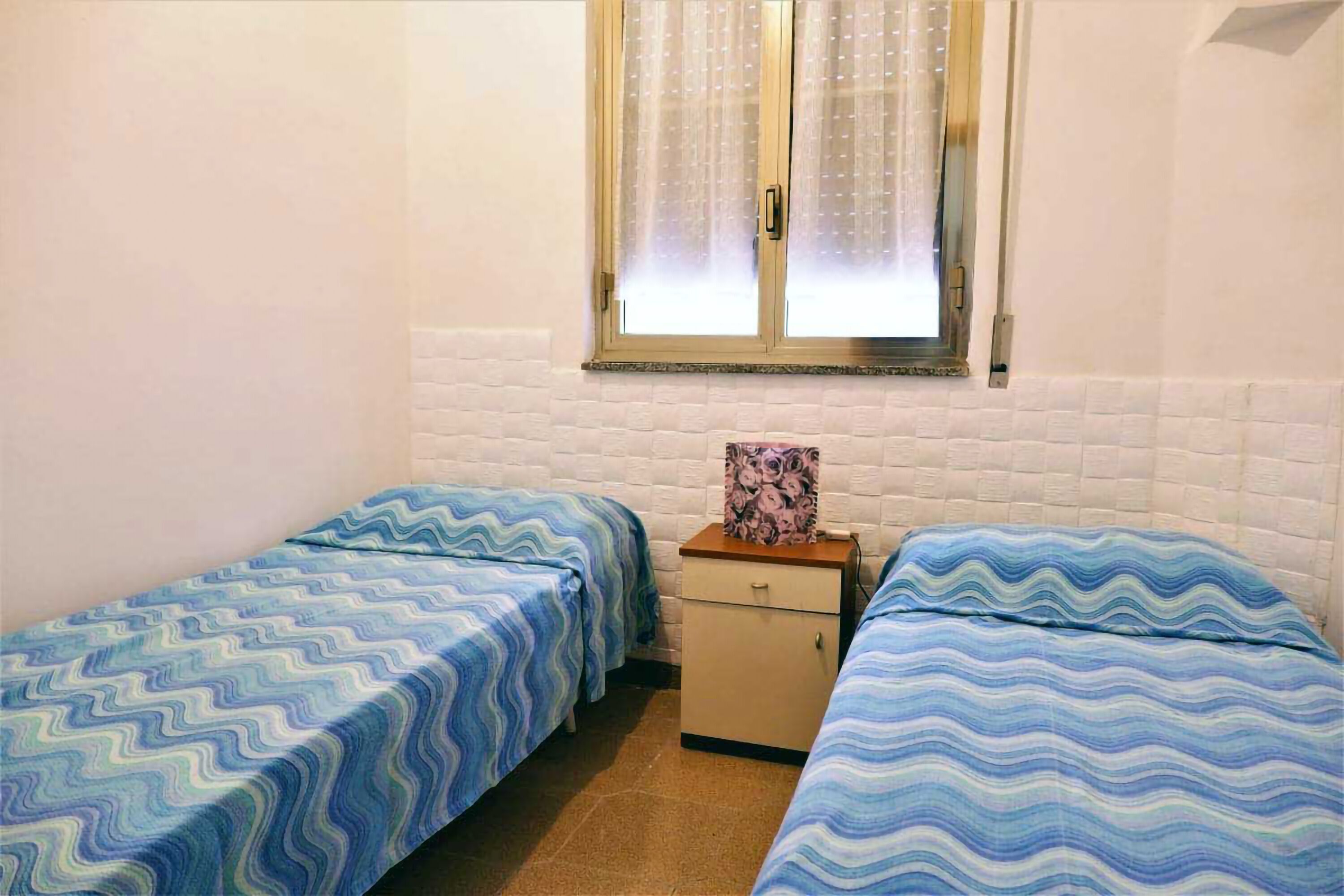 A small, single-room accommodation with two single beds, a small wooden cabinet, and a window with a curtain.