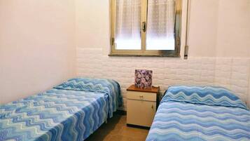 A small, single-room accommodation with two single beds, a small wooden cabinet, and a window with a curtain.