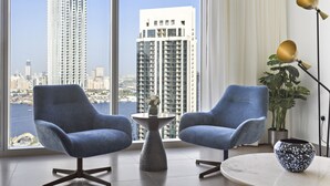 A modern room with two blue armchairs, a small round table with a vase, a floor lamp, and a view of tall buildings through large windows. - WelHome - Luxe Apartment With Incredible View on Dubai Creek (Dubai)