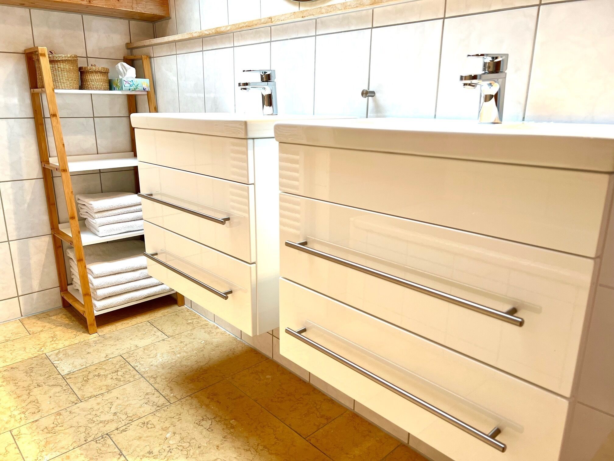 Ein Badezimmer mit weißen Fliesen, einem Holzregal mit Handtüchern und einem Waschbecken mit chromfarbenen Wasserhahn.