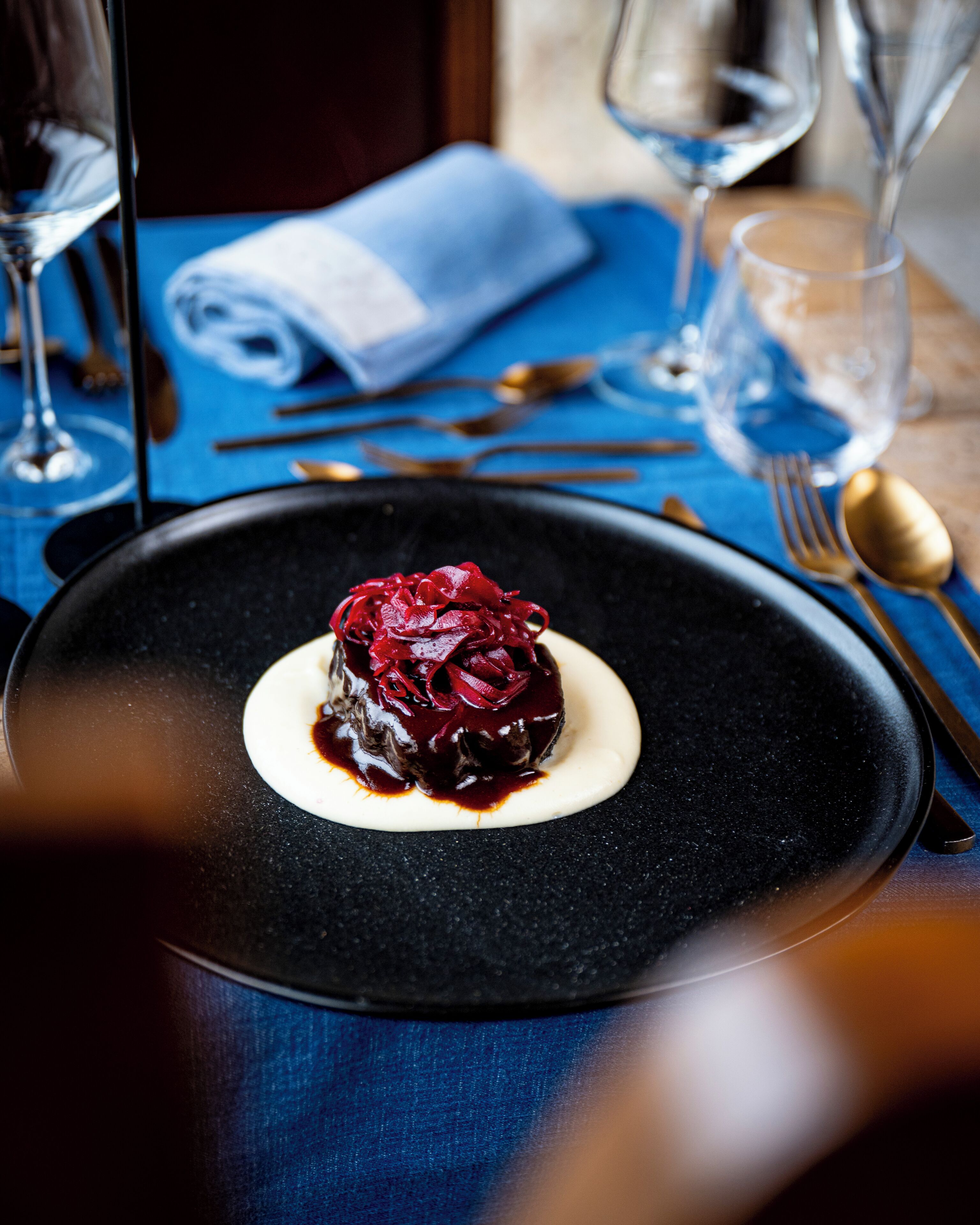 A dessert on a black plate with a blue tablecloth, accompanied by a folded napkin, cutlery, and wine glasses.