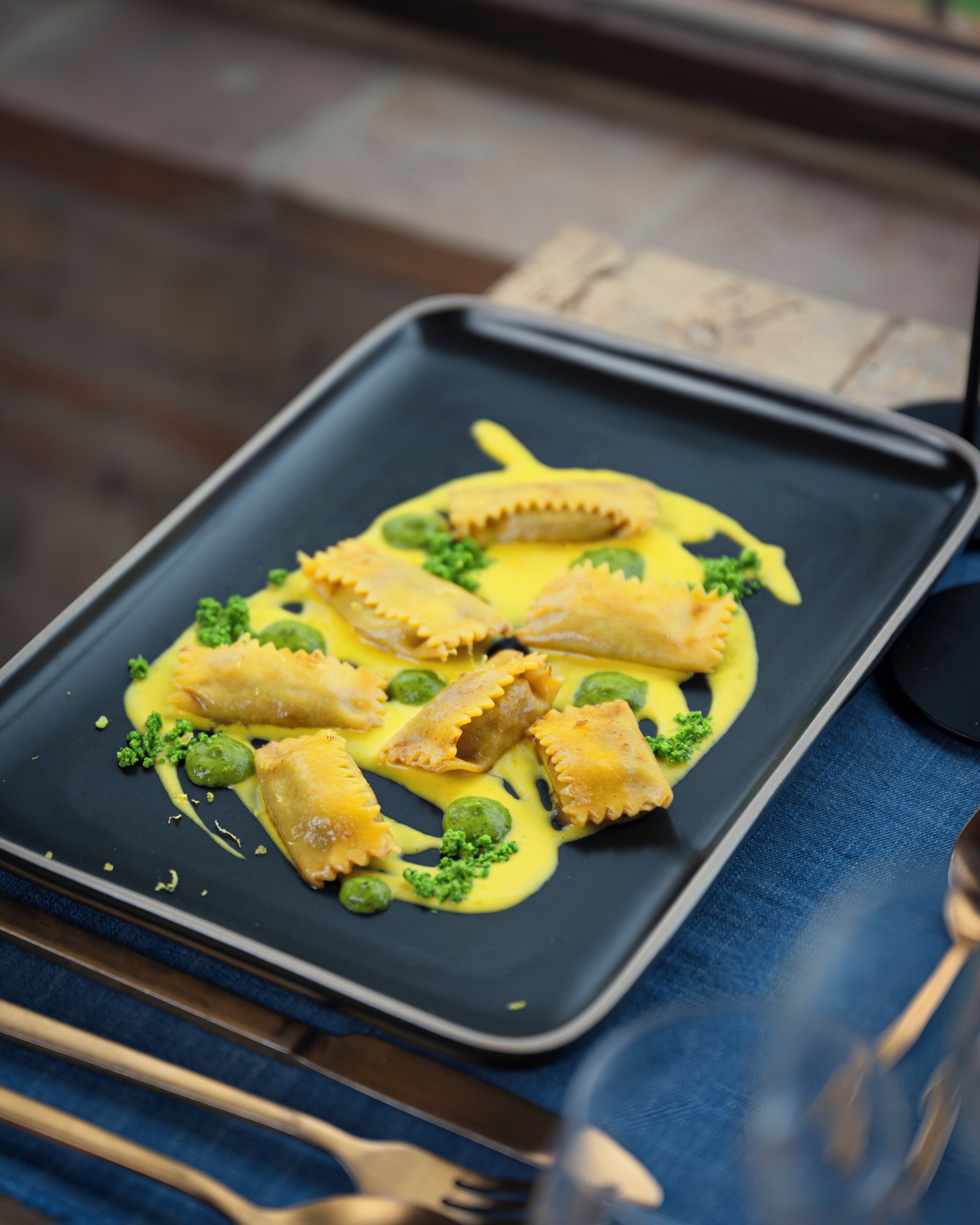 Un piatto nero rettangolare con ravioli, piselli verdi e una salsa gialla, adagiato su una tovaglia blu.