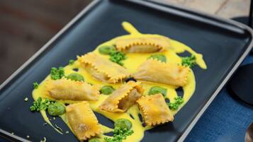 A rectangular black plate with ravioli, green peas, and a yellow sauce, placed on a blue tablecloth.