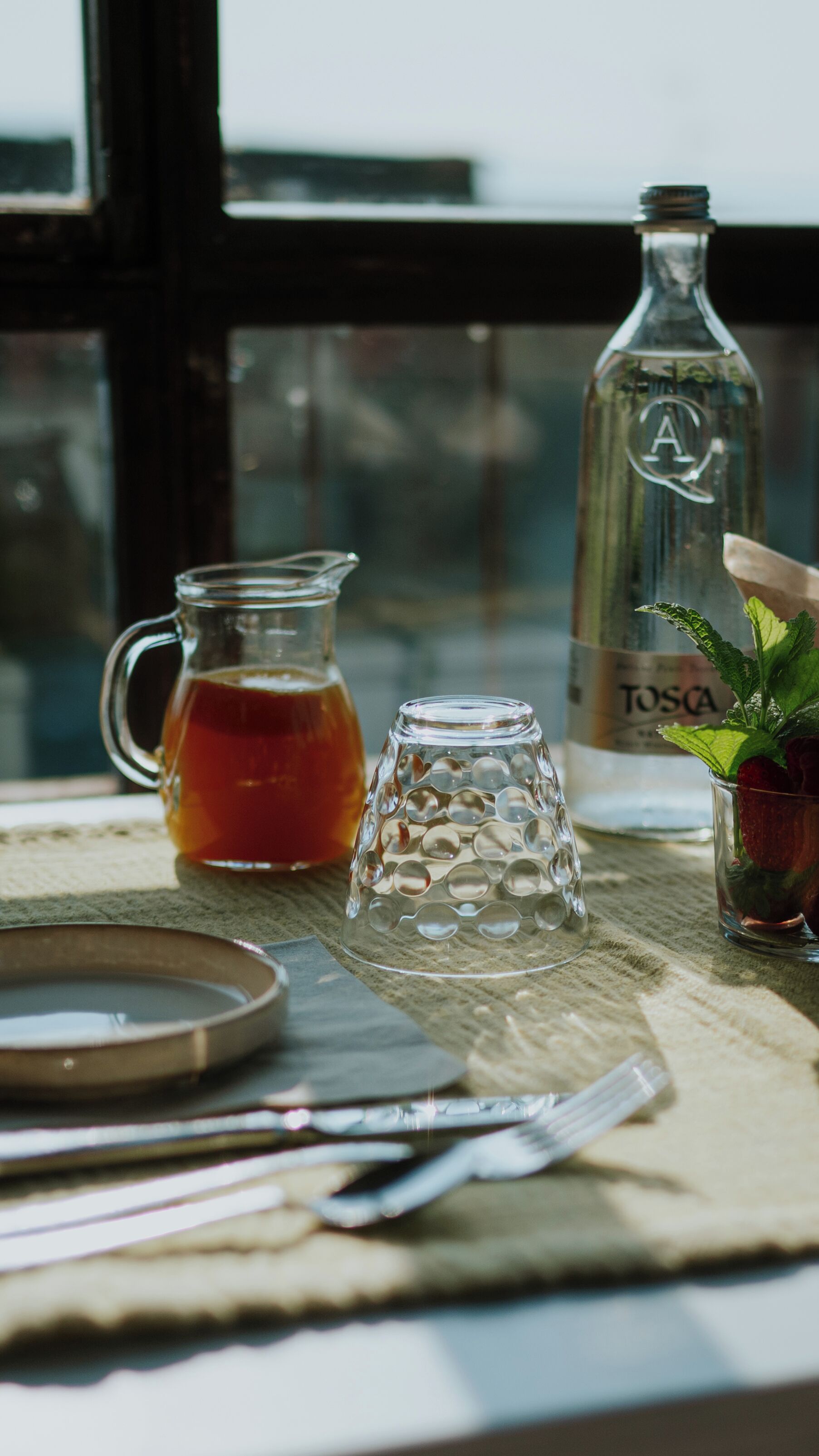 Una mesa con un jarro de vidrio, una jarra, un vaso decorado con una ramita de menta y una botella etiquetada 'Tosca'.