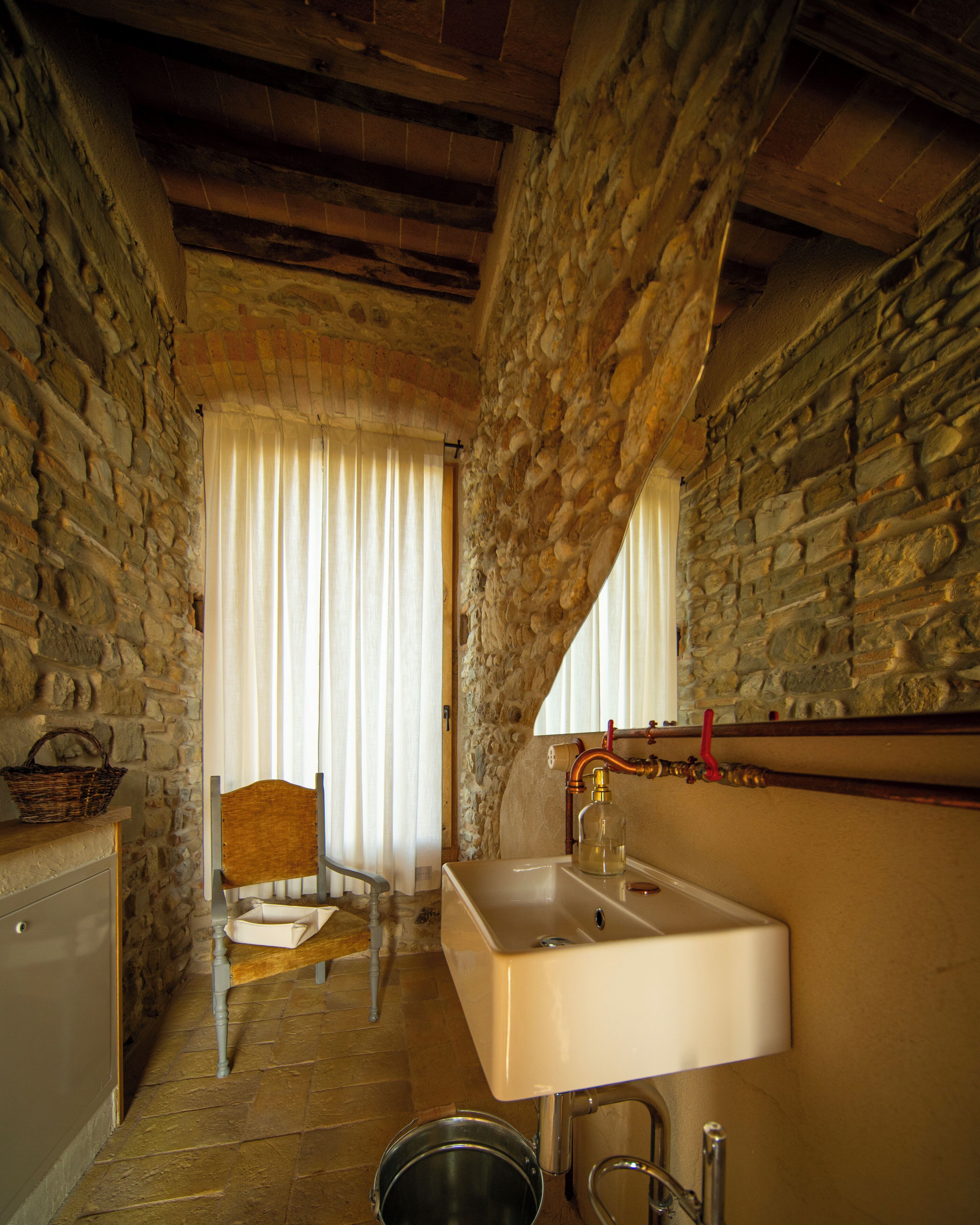 A narrow stone-walled bathroom with a wooden chair, a white sink, and a metal bucket.