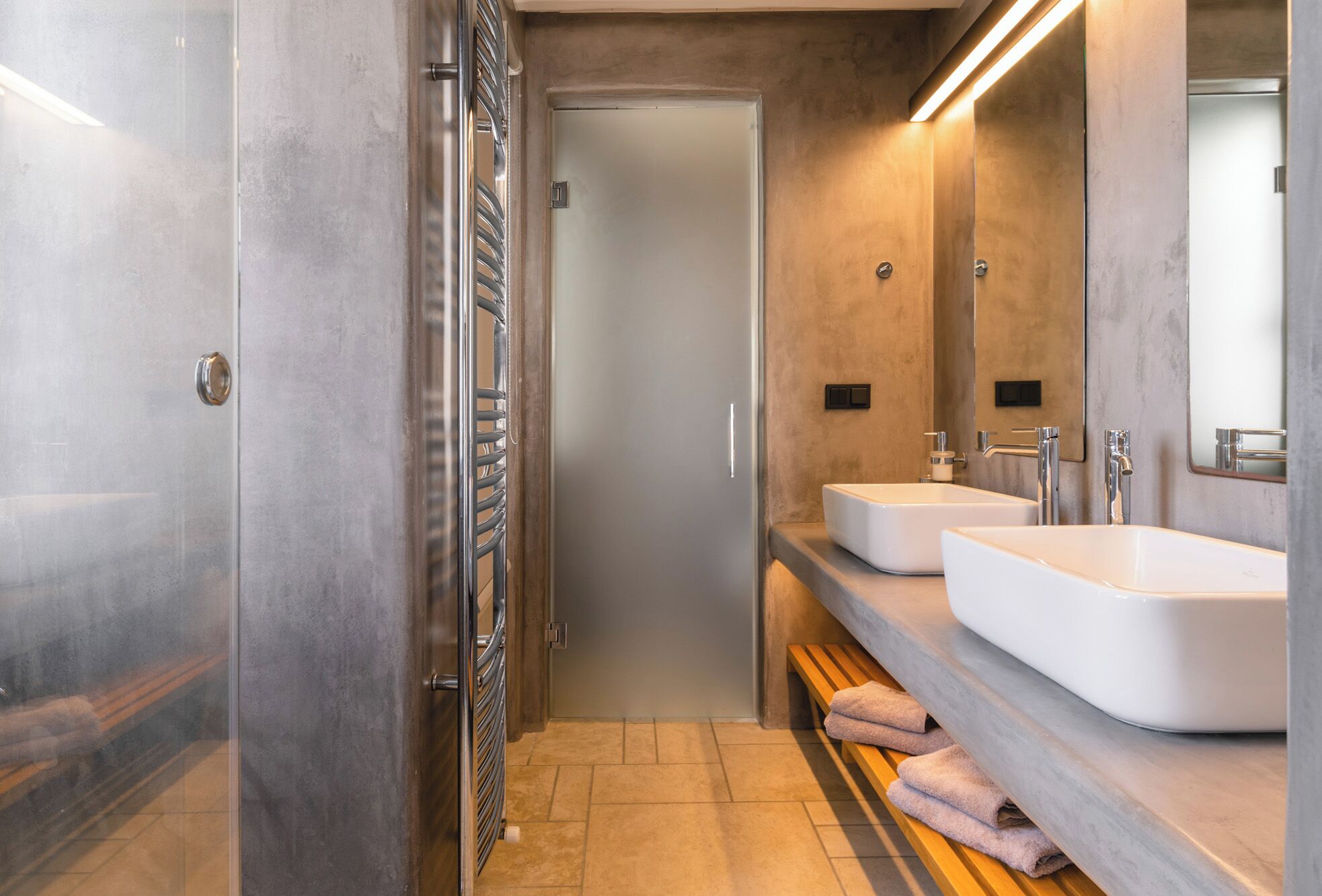 A modern bathroom with a glass shower enclosure, two sinks, and a bench with towels.