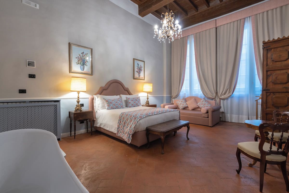 A bedroom with a bed, bedside tables, a sofa, a chair, and a chandelier.