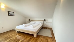 Una cama bien hecha con sábanas y toallas blancas, una mesita de noche con lámpara y una pequeña alfombra sobre el suelo de madera.