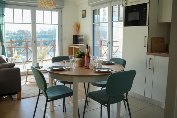 A dining area with a round table set for four, surrounded by green chairs. There is a television on a shelf above the table, and a microwave above the cabinets to the right.