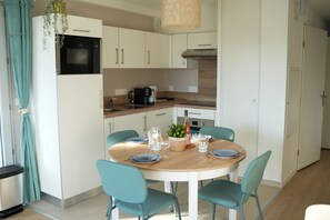 A modern kitchen with white cabinets, a built-in microwave, and a coffee maker. There is a dining table with chairs and a potted plant.