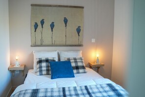 A neatly made bed with a blue pillow, flanked by two bedside tables with lamps, and a wall-mounted painting of birds.