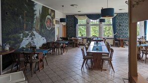 A dining area with multiple tables and chairs, large windows with a view of greenery, and a mural on the wall. - Wanderhotel Steinerne Renne (Wernigerode)