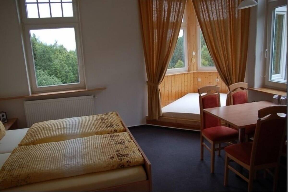 A room with a bed, a table, and chairs. There are windows with curtains.