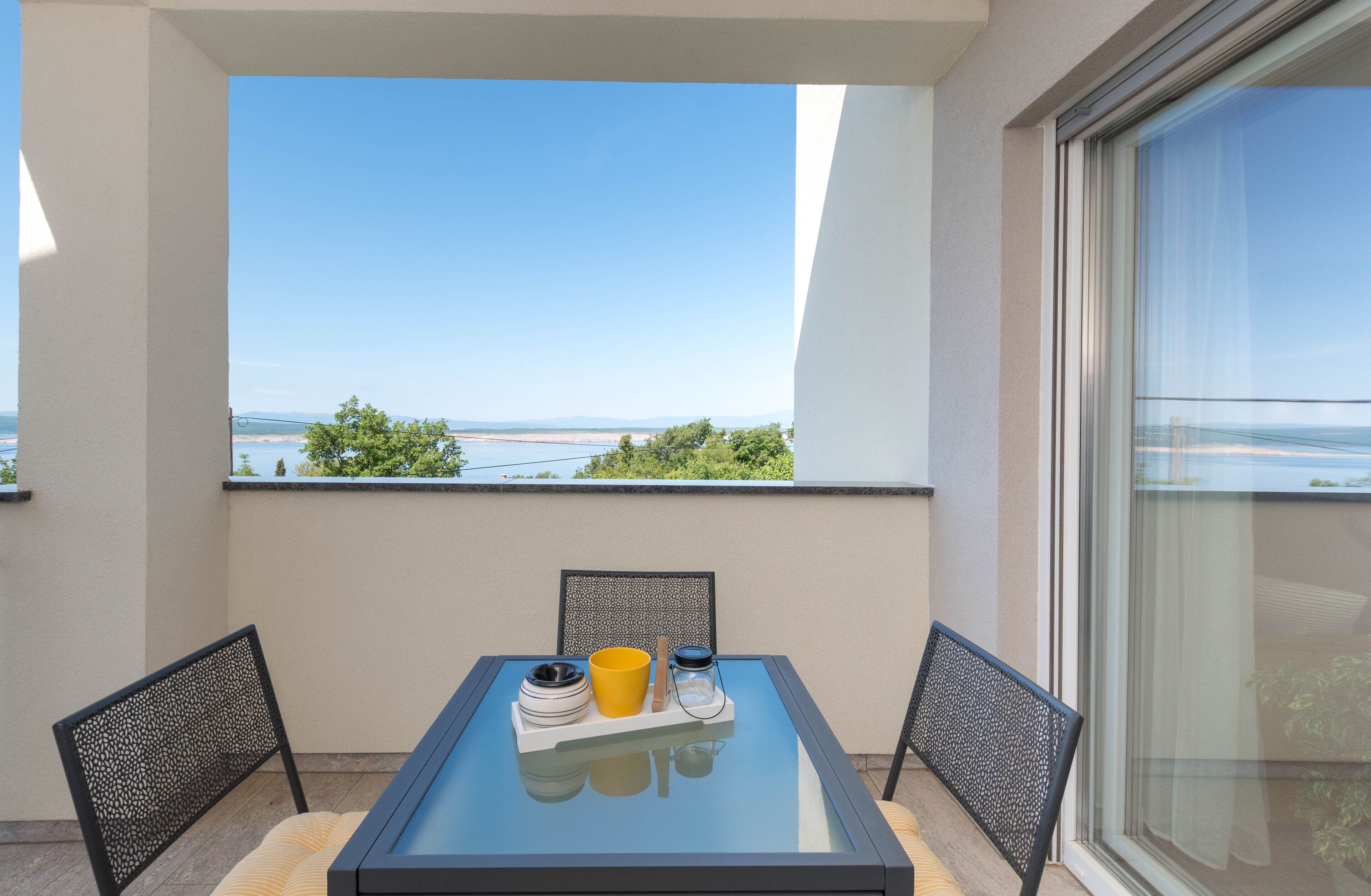 Un balcón con una mesa de vidrio, dos sillas y vista al mar.