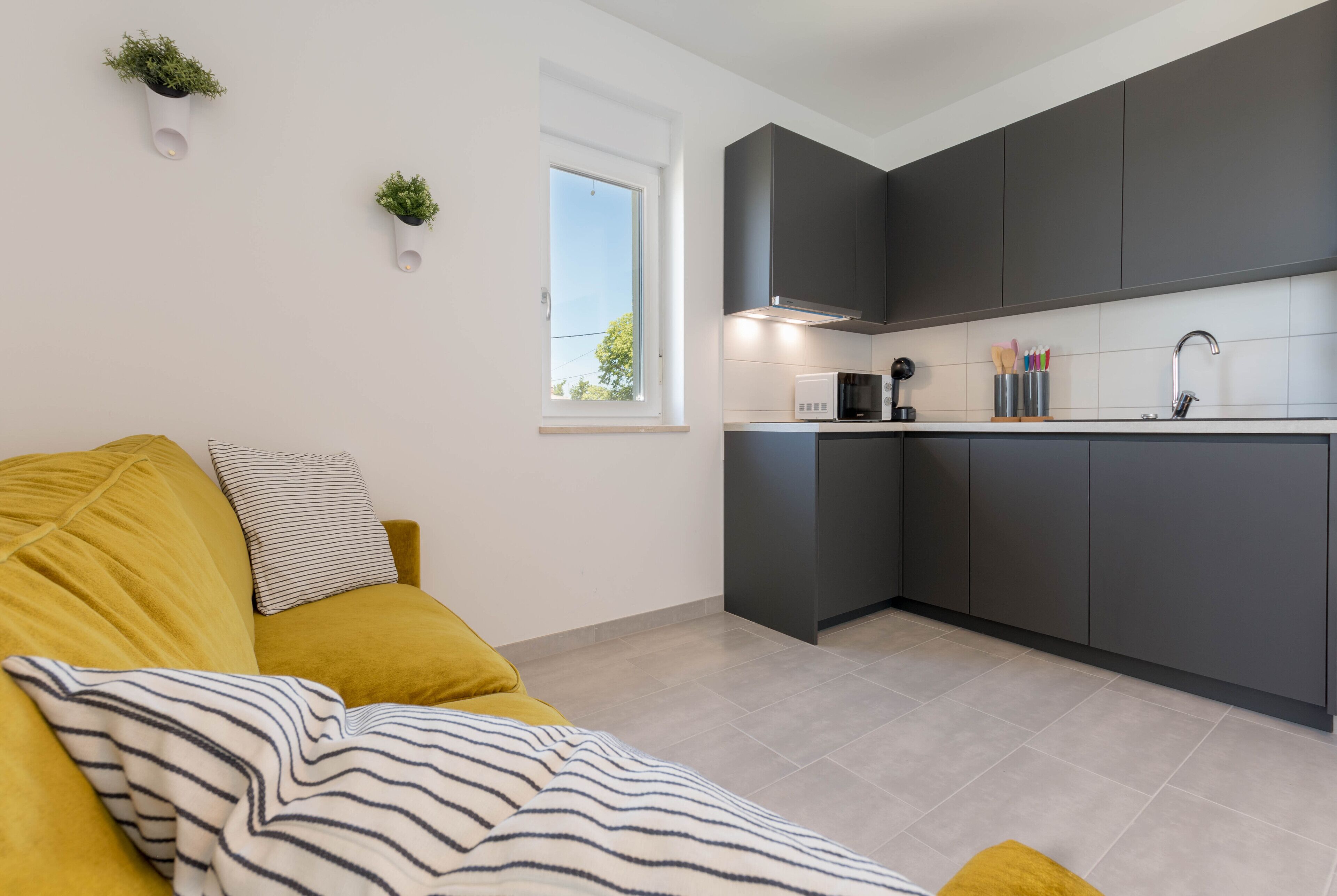Una cocina moderna con armarios oscuros, microondas y pileta. Se observa una ventana que proporciona luz natural, y un sofá amarillo con almohadones a rayas al frente.